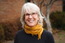 Betsy Humphreys is a woman is midlength white hair, tortoise shell glasses, and a bright yellow scarf