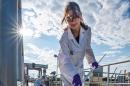 Woman in white lab coat samples liquid outdoors with bright sun over her shoulder